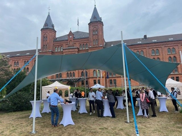 Eine Gruppe von Menschen steht unter einem Zelt vor einem großen Gebäude.