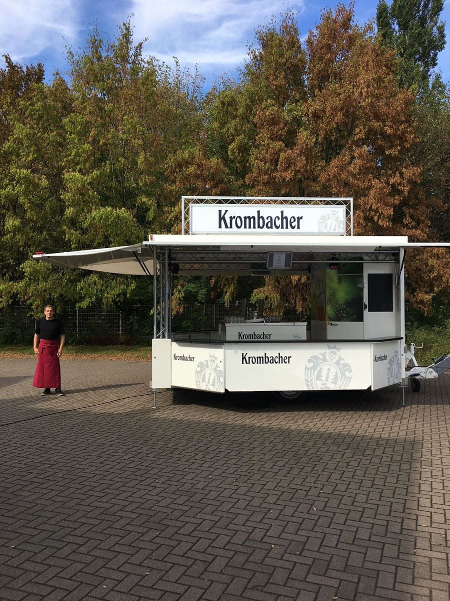 Ein Mann steht vor einem Imbisswagen, auf dem „Krambacher“ steht.