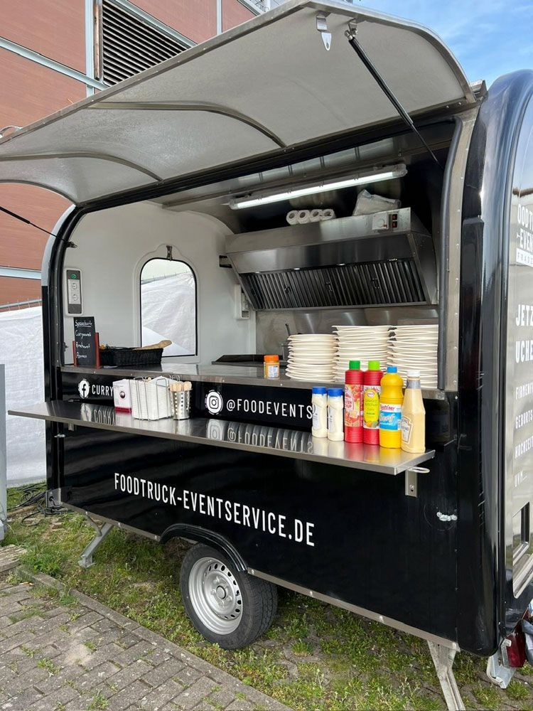 Ein schwarzer Imbisswagen steht mit offener Tür vor einem Gebäude.