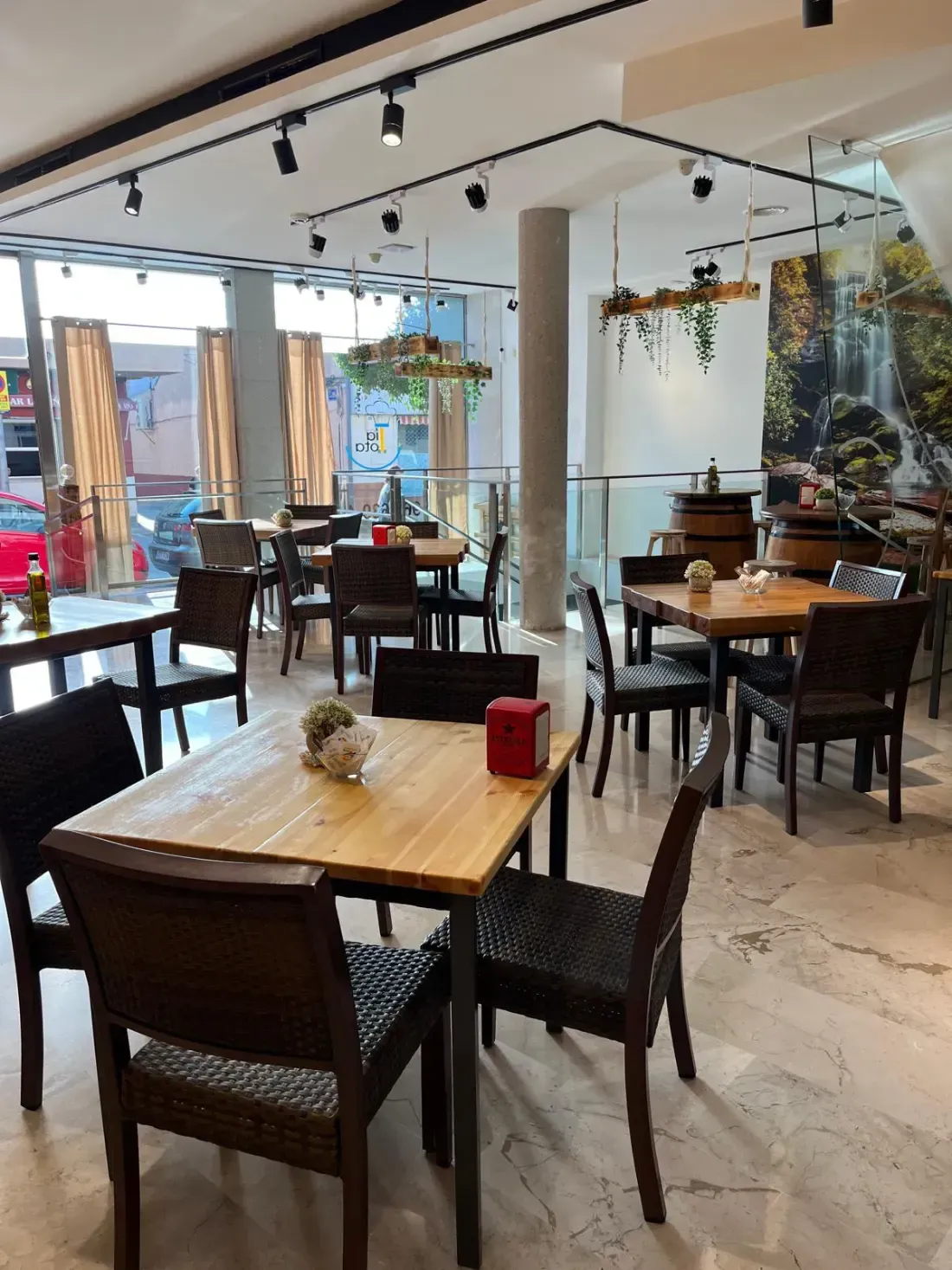 Interior de restaurante con mesas y sillas, luz natural y un mural de una cascada.