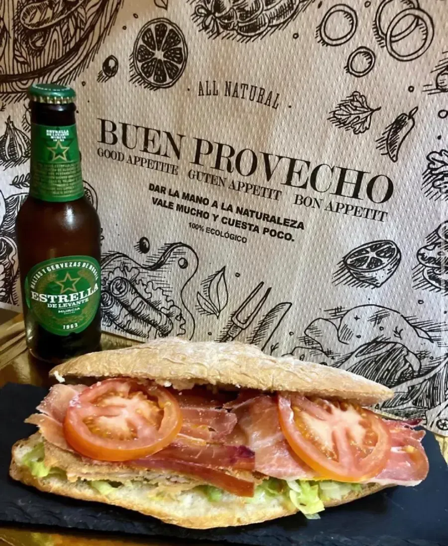 Sándwich de tomate y jamón junto a una botella de cerveza. Texto