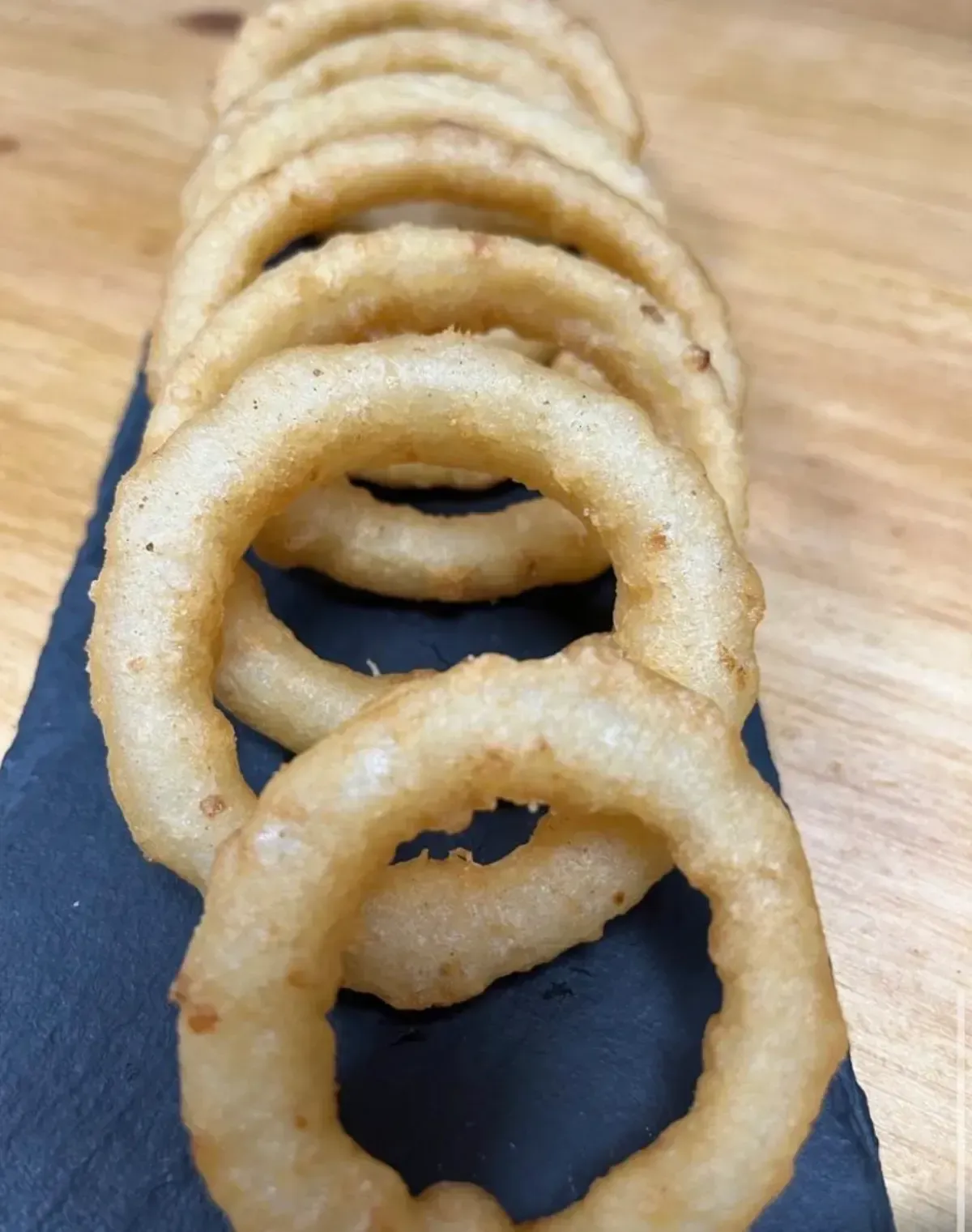 Aros de cebolla fritos dorados sobre una pizarra oscura, mesa de madera al fondo.