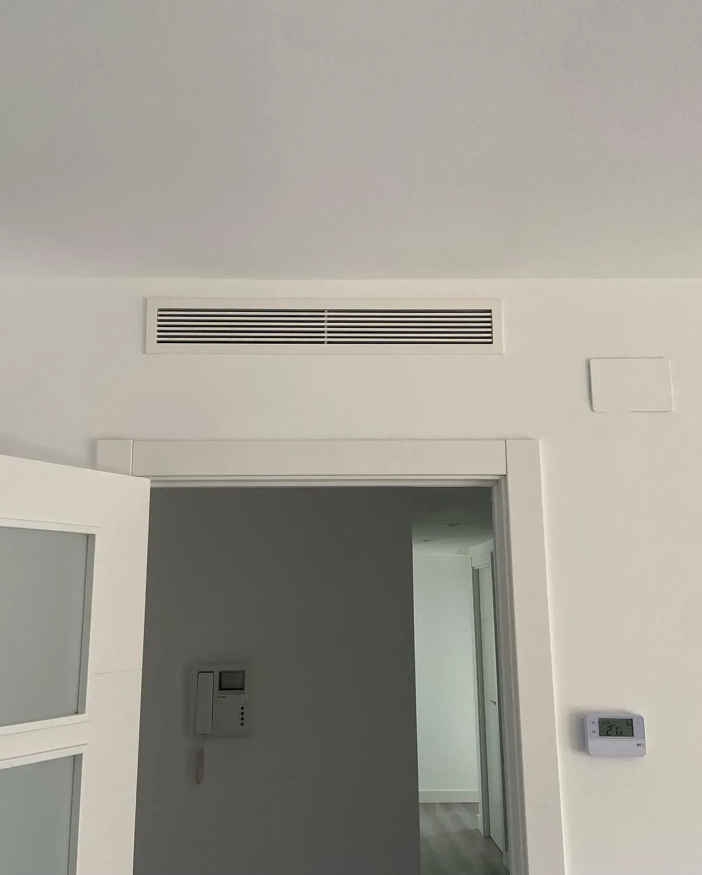 Entrada interior con una puerta blanca parcialmente abierta, una rejilla de ventilación en la parte superior de la pared