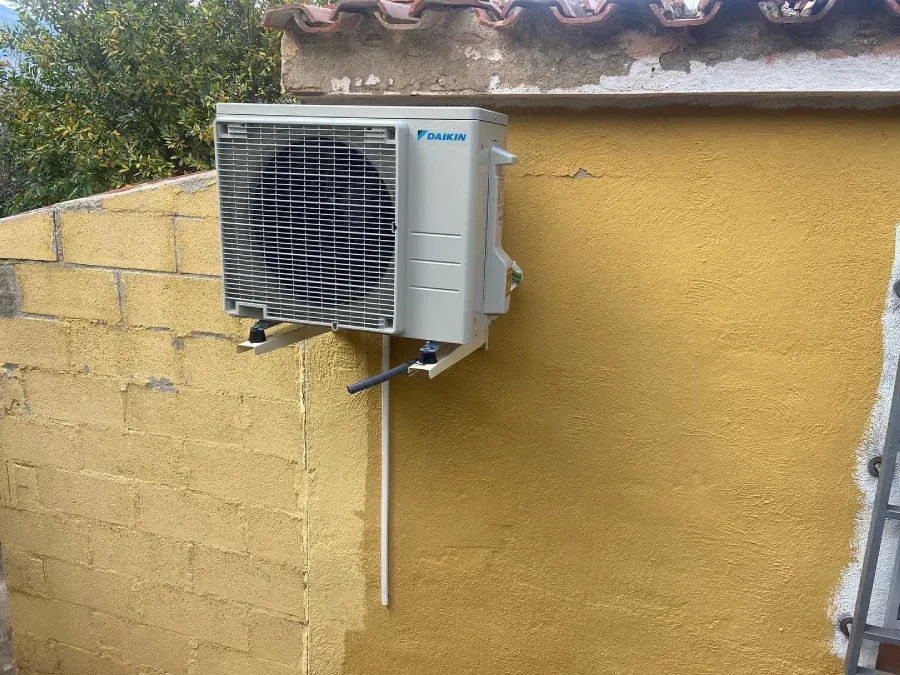 Unidad exterior de aire acondicionado instalada en una pared de estuco amarillo, con tuberías y soporte visibles