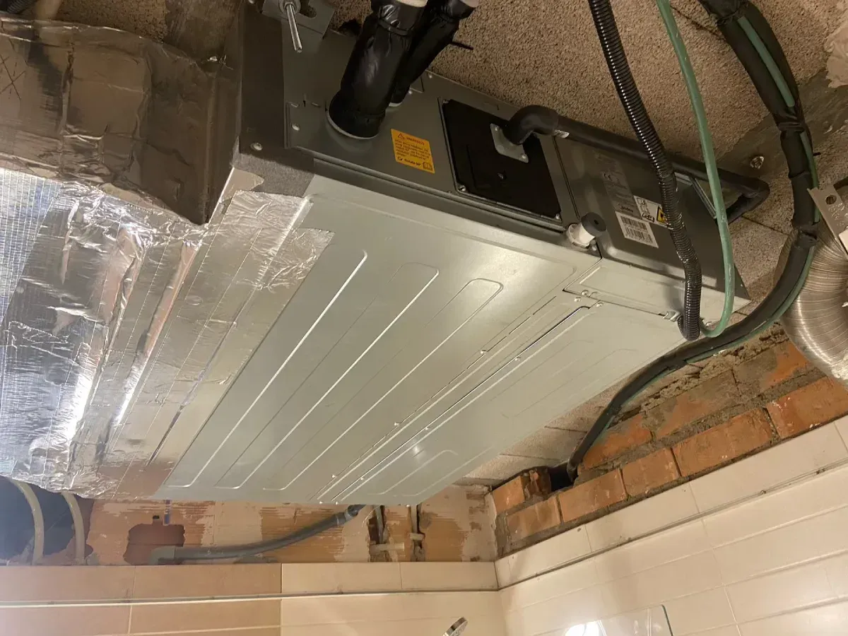 Parte inferior de una unidad metálica de climatización instalada sobre una pared de azulejos blancos