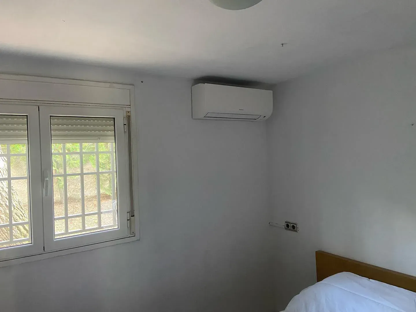 Dormitorio blanco vacío con una ventana, aire acondicionado de pared y el borde de la cama a la vista
