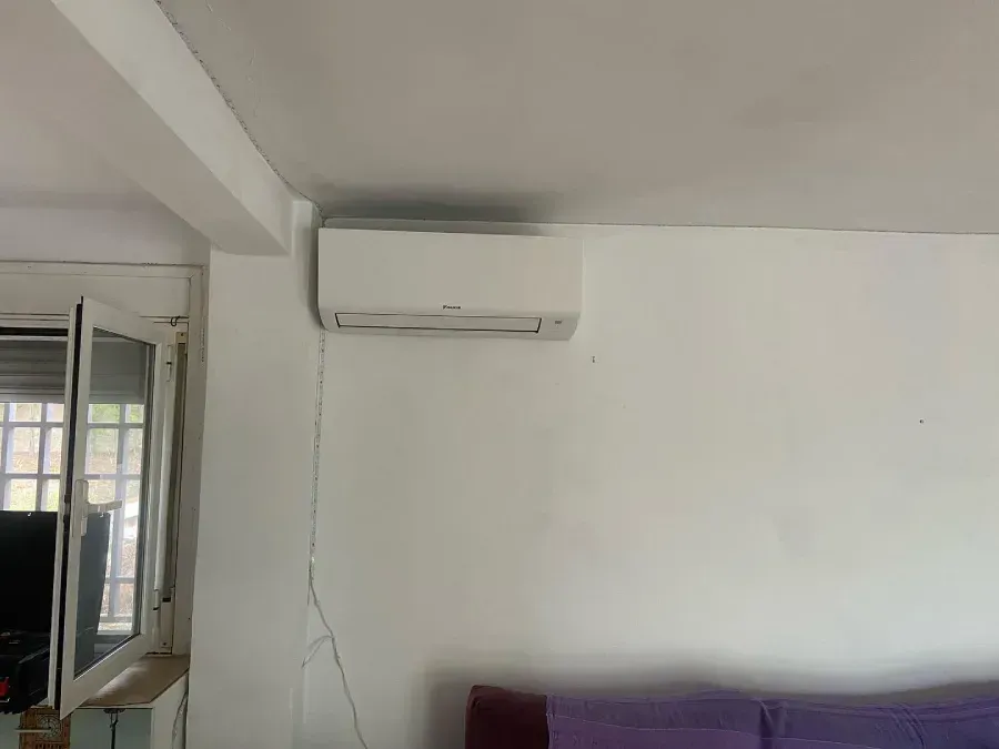 Habitación blanca con un aire acondicionado montado en la pared, una ventana a la izquierda y un sofá morado debajo.