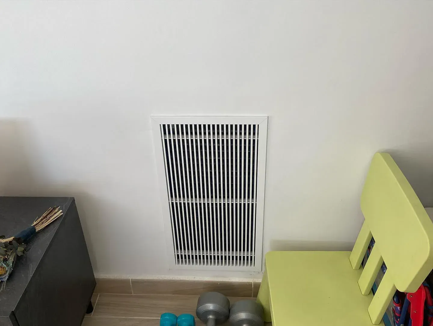 Esquina de una habitación pequeña con una rejilla de ventilación en la pared, dos pesas rusas grises