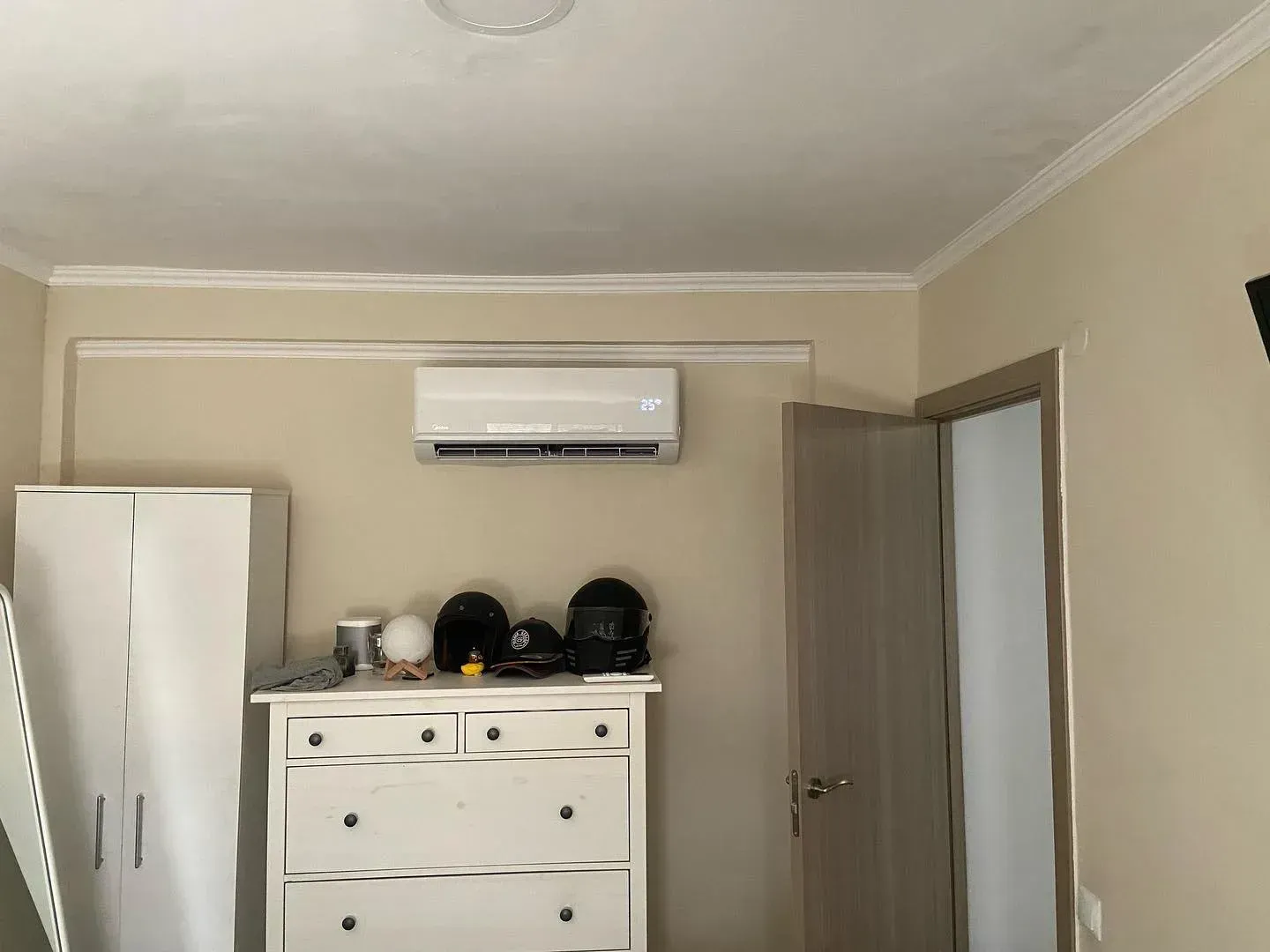 Una habitación beige con una cómoda blanca, un armario alto, un aire acondicionado de pared y una puerta.