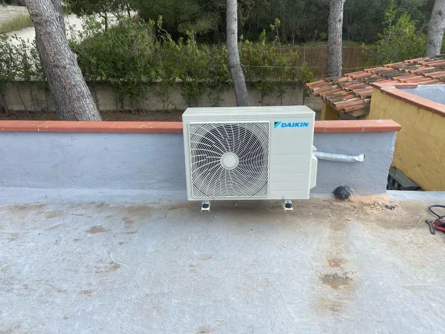 Unidad de aire acondicionado exterior en una azotea, junto a un muro bajo y árboles, con una tubería de desagüe y cableado.