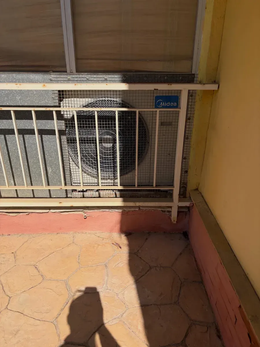 Unidad exterior de aire acondicionado detrás de una barandilla metálica blanca, junto a una pared amarilla