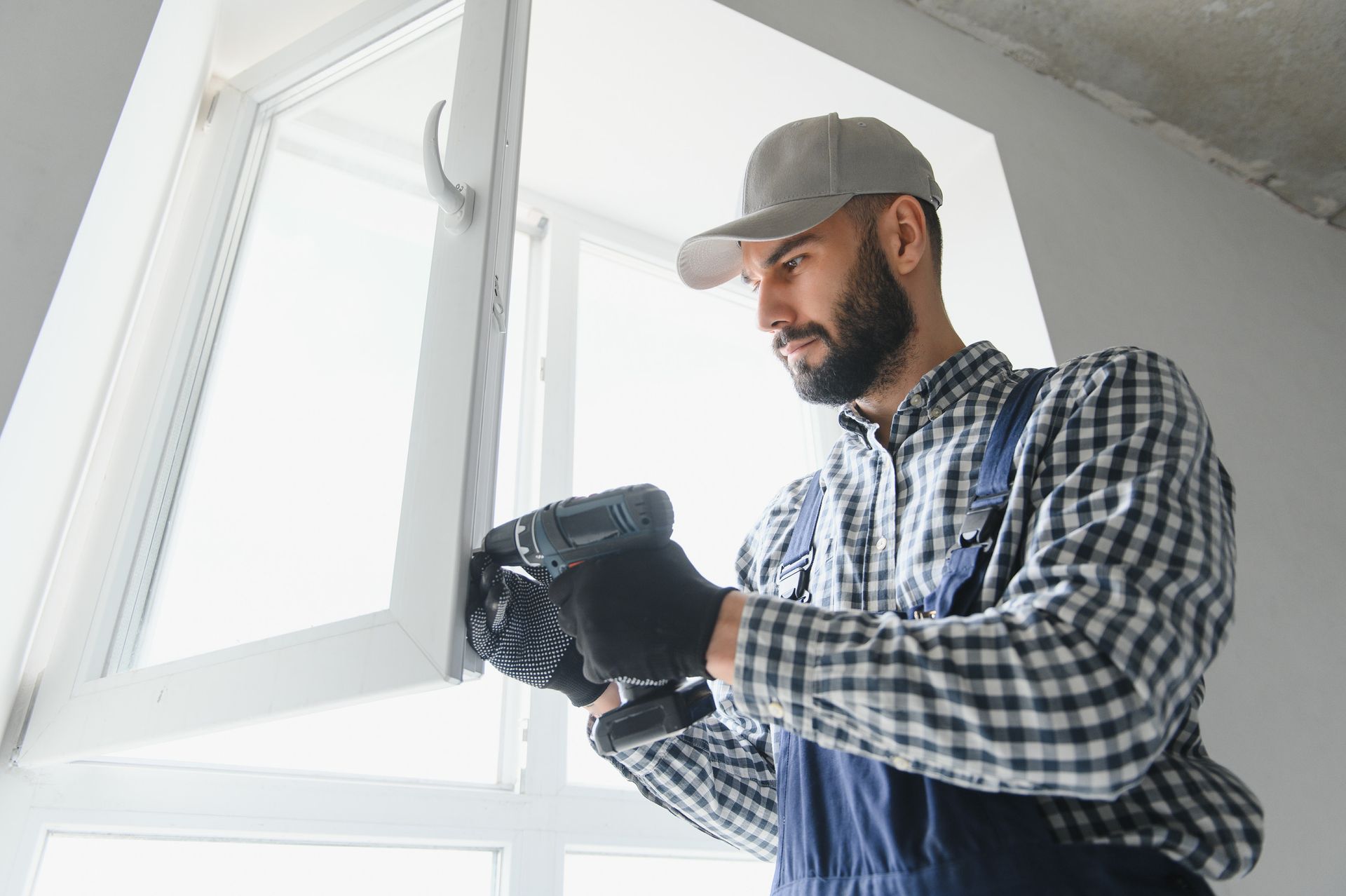 Hombre con mono azul y gorra usando un taladro para instalar una ventana en una habitación de paredes blancas.