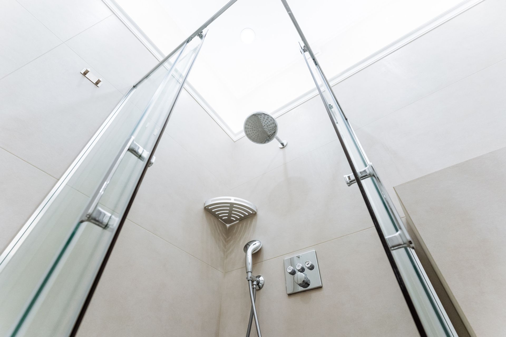 Cabina de ducha con puertas de vidrio, cabezal de ducha tipo lluvia y cabezal de ducha de mano contra una pared de tono neutro.