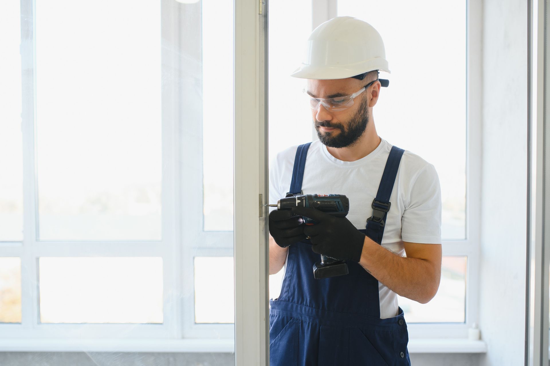 Un trabajador de la construcción con mono perfora el marco de una puerta cerca de una ventana.