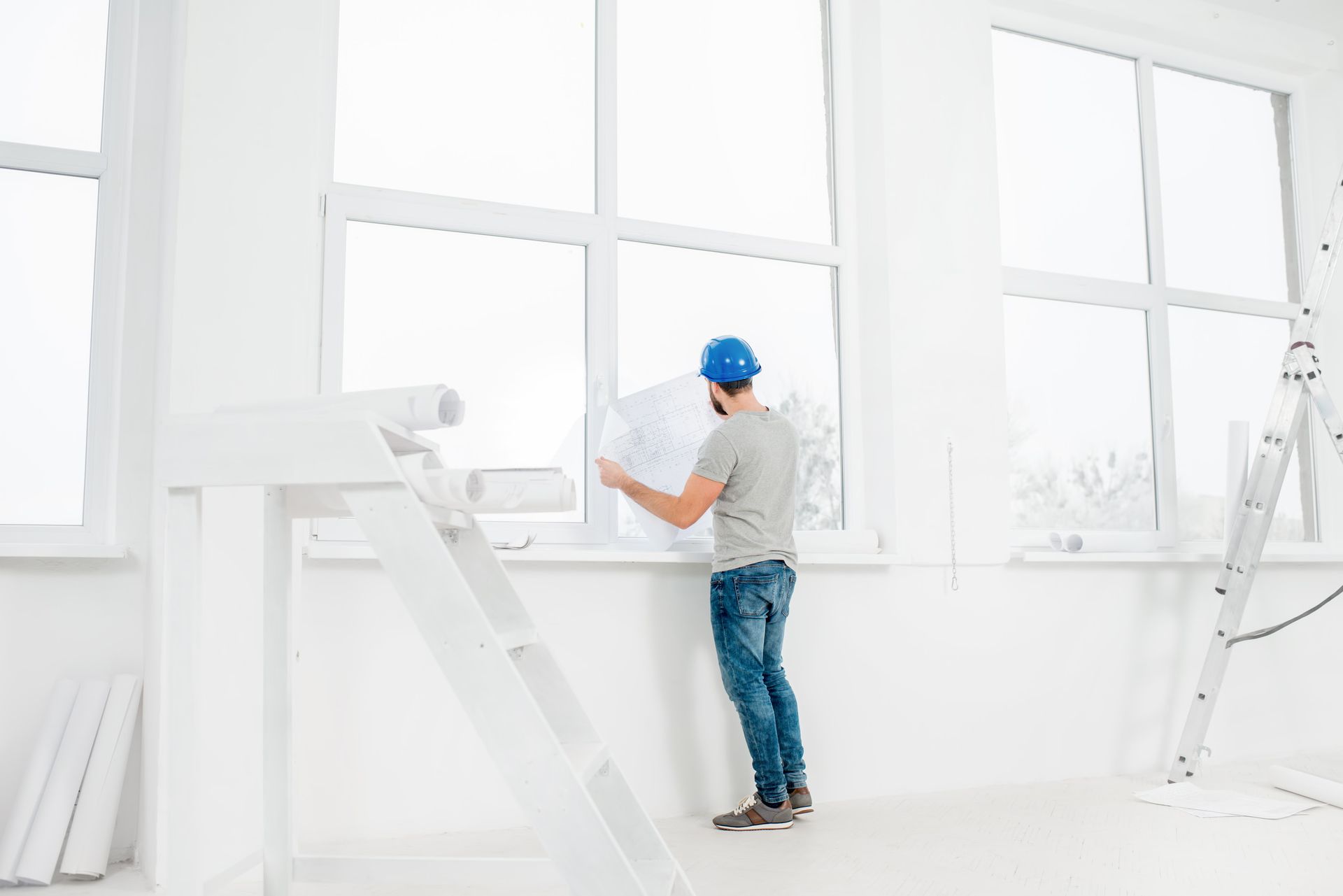 Hombre con casco azul estudiando planos junto a una ventana en una habitación blanca y luminosa.