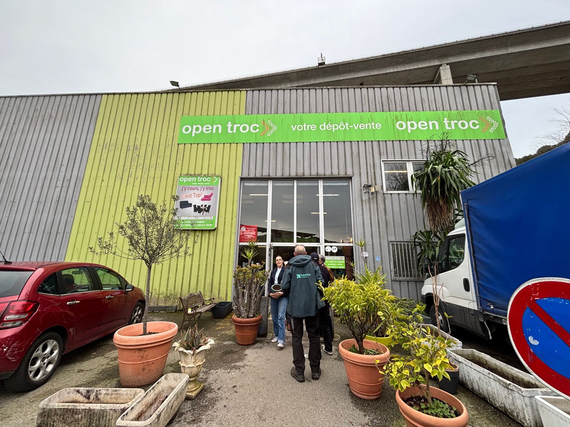 Vue extérieure d'un magasin Open troc avec des clients entrants. Façade vert, gris et jaune, plantes et véhicules.