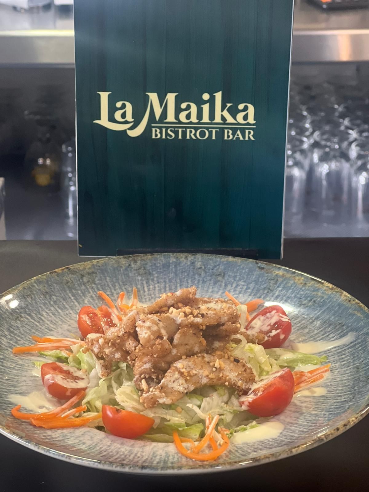 Ensalada con pollo frito, tomates y zanahorias frente a un cartel de La Maika Bistro Bar.