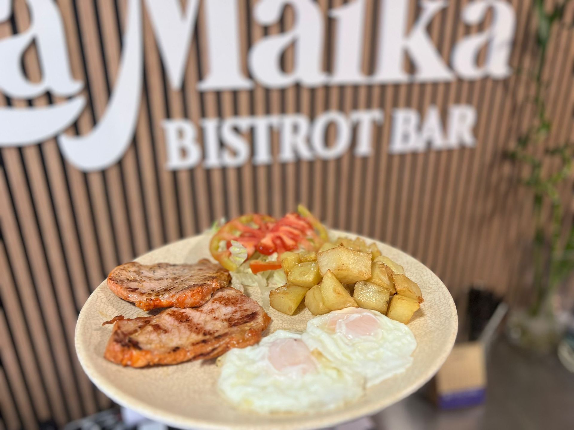 Plato de comida: cerdo a la parrilla, huevos fritos, patatas y tomates delante del cartel de un bar bistró.