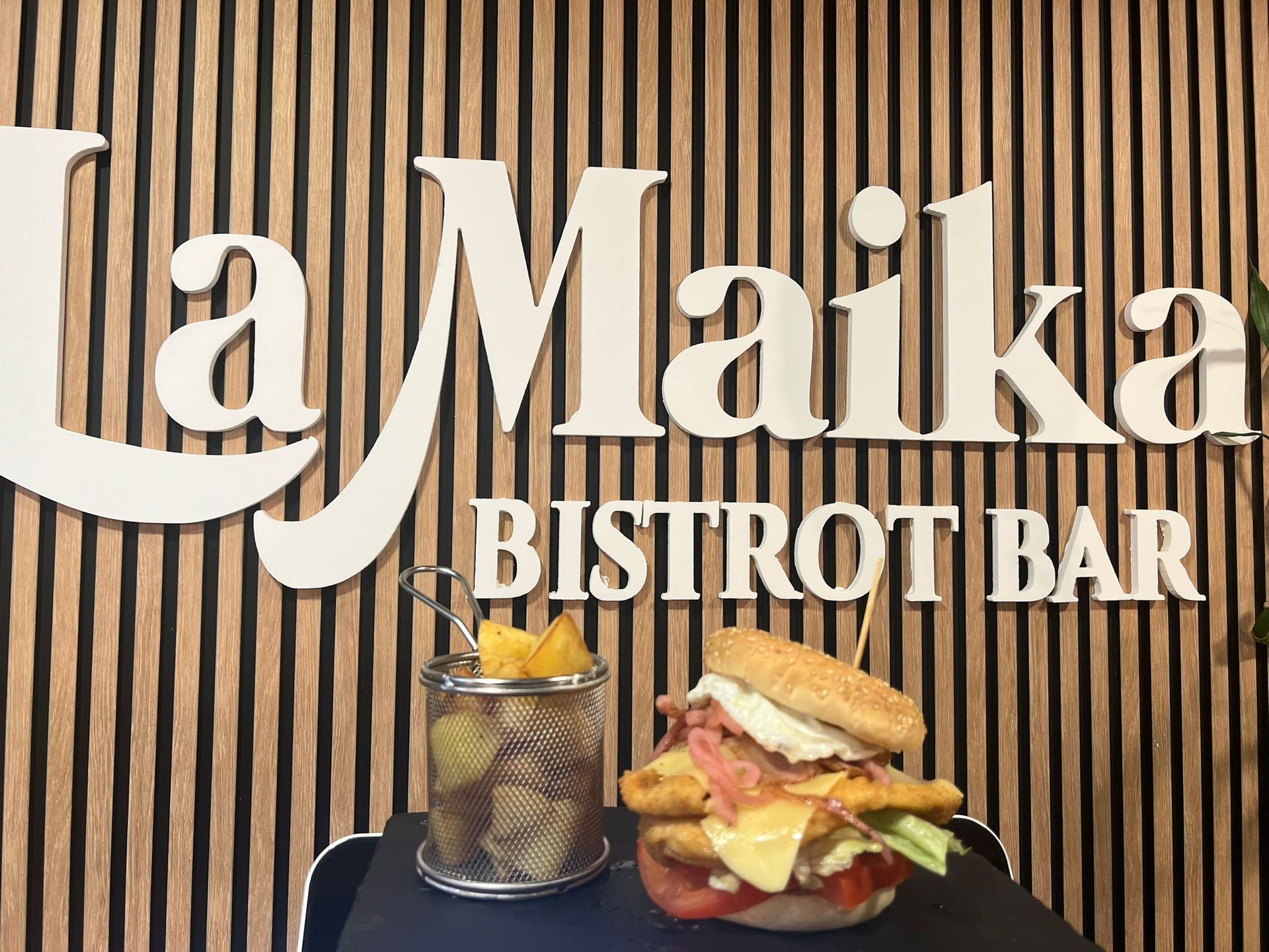 Sándwich y patatas fritas frente al cartel de La Maika Bistro Bar sobre fondo de panel de madera.