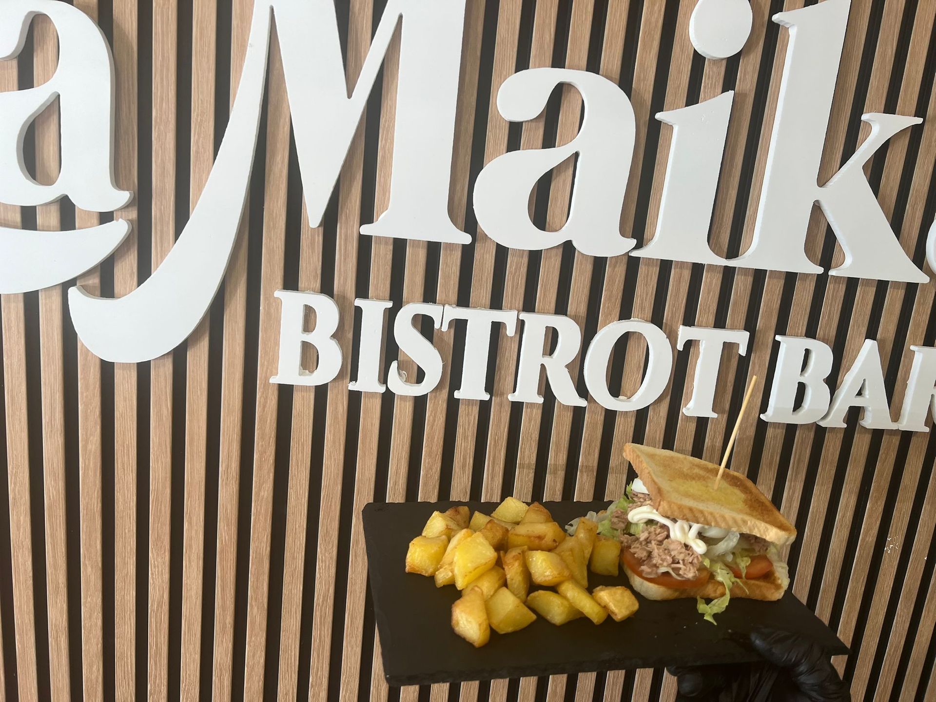 Una mano sosteniendo un plato con un sándwich y patatas, delante del cartel 