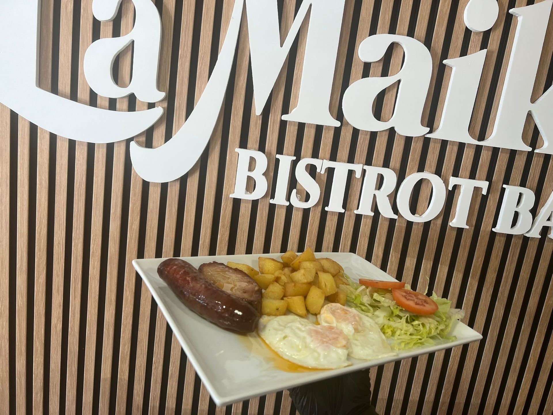 Plato de comida con salchichas, patatas y huevos delante del logotipo de un restaurante.