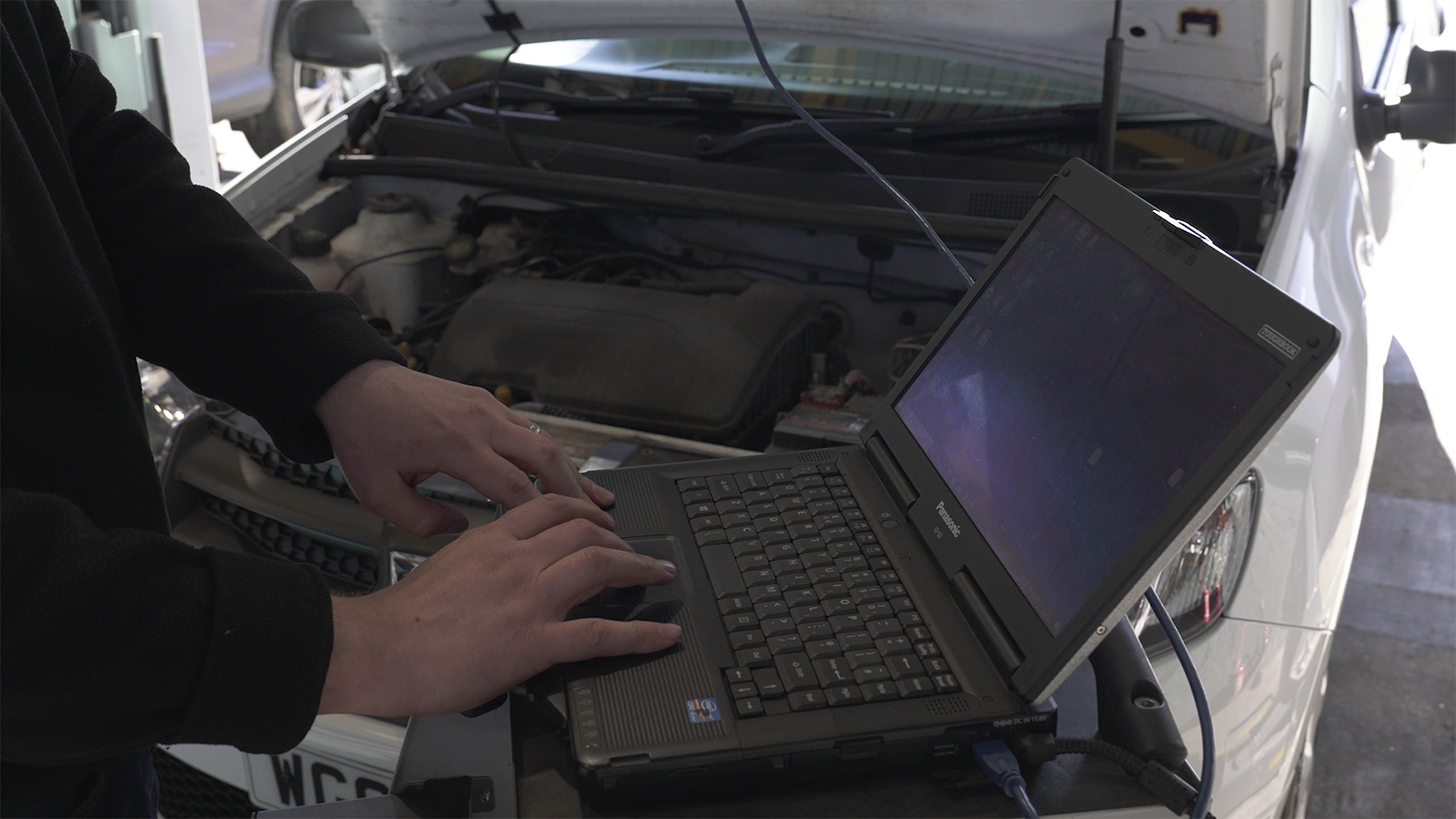 A person is typing on a laptop in front of a car.