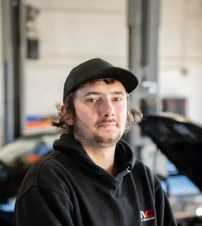 A man wearing a hat and a hoodie is sitting in front of a car with the hood open.