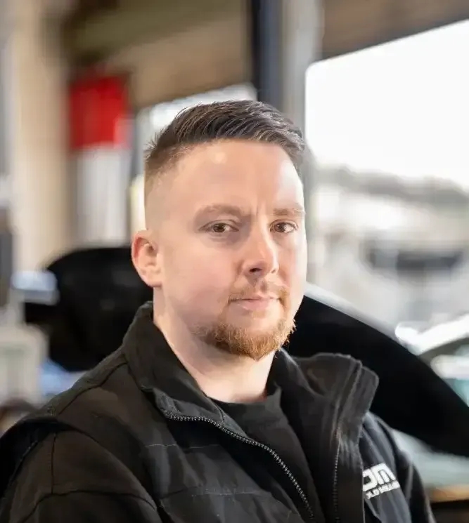 A man with a beard is sitting in front of a car and looking at the camera.