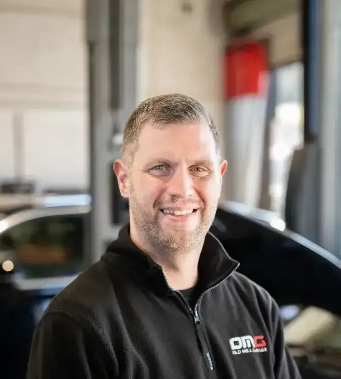 A man in a black jacket is smiling in front of a car.