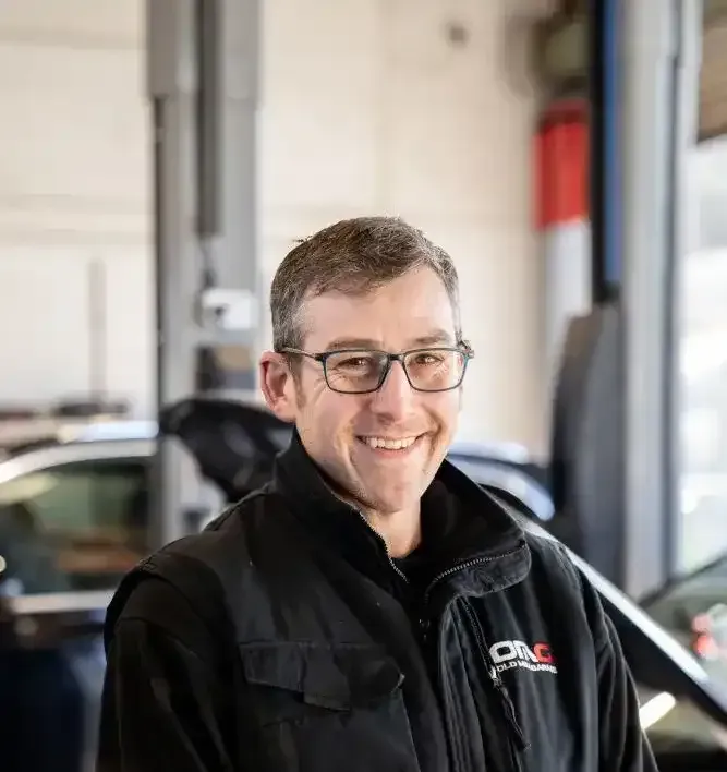 A man wearing glasses is smiling in front of a car in a garage.