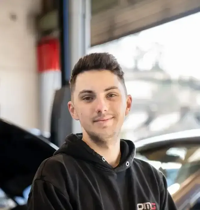 A young man wearing a black hoodie is sitting in front of a car in a garage.