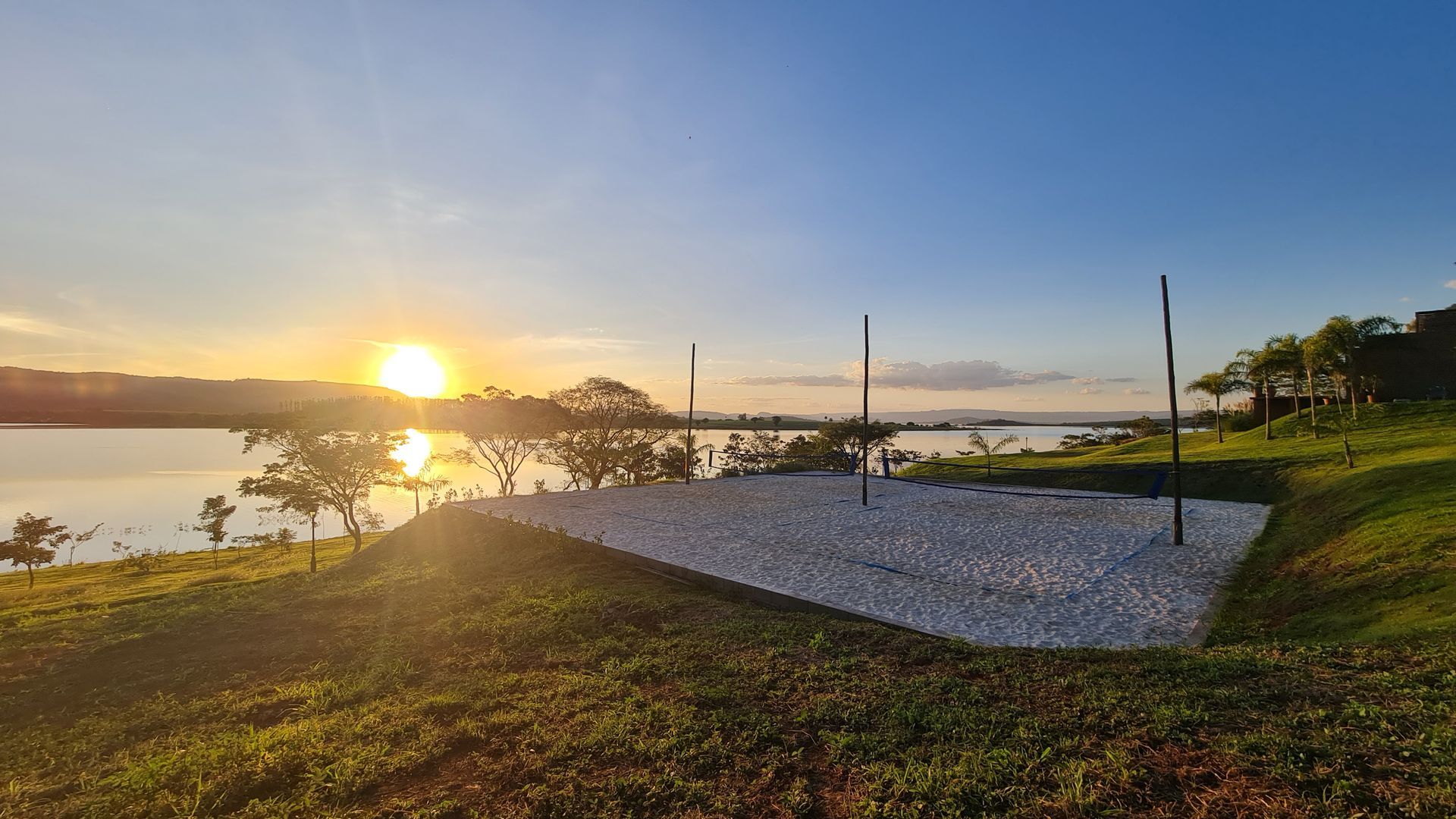 Uma quadra de vôlei de praia à beira de um lago ao pôr do sol. O céu está azul com um sol brilhante. A quadra tem areia branca.