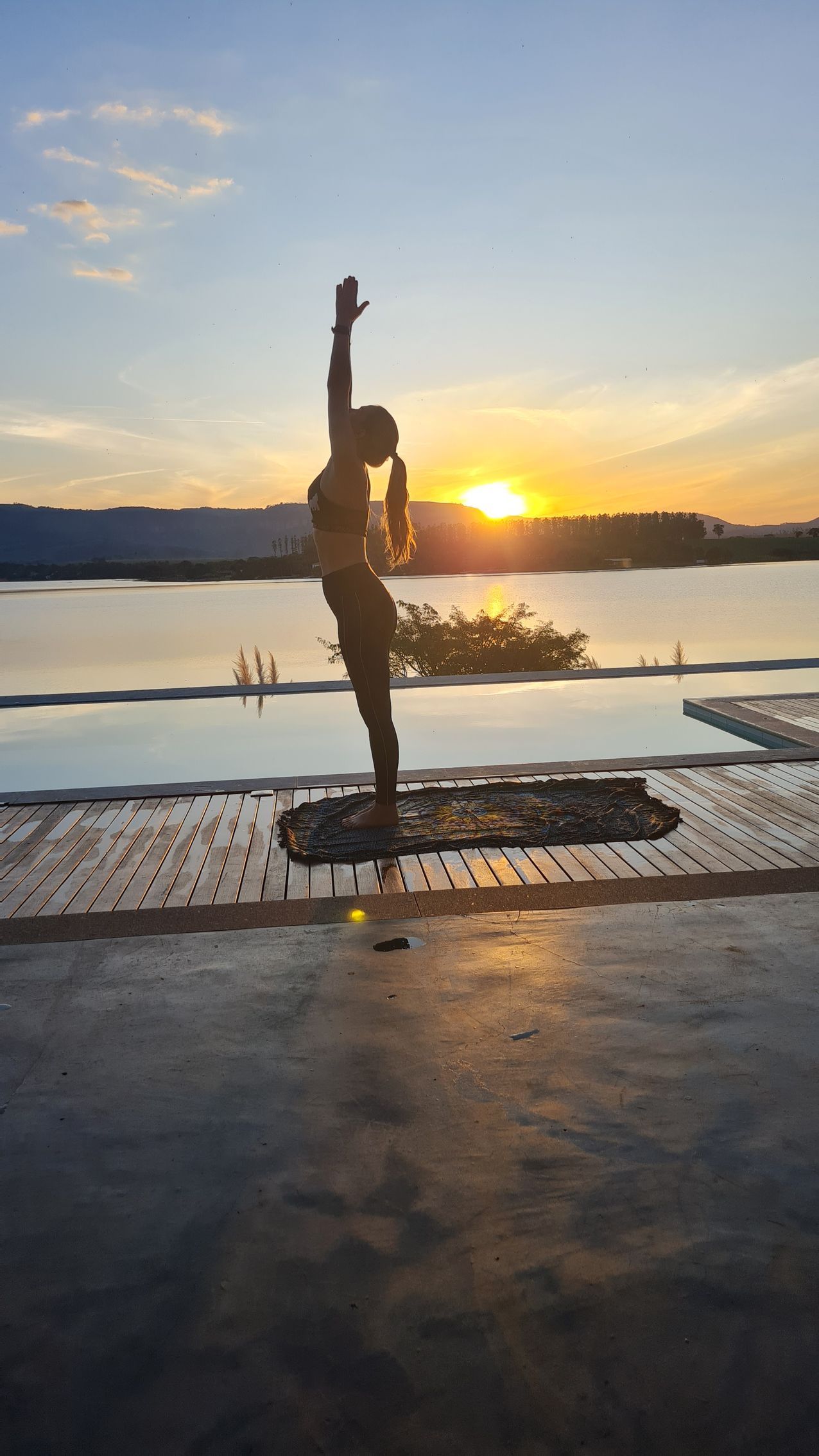 Mulher em pose de ioga ao pôr do sol, com os braços levantados, em pé sobre um tapete perto de piscinas com níveis e o sol refletindo na água.