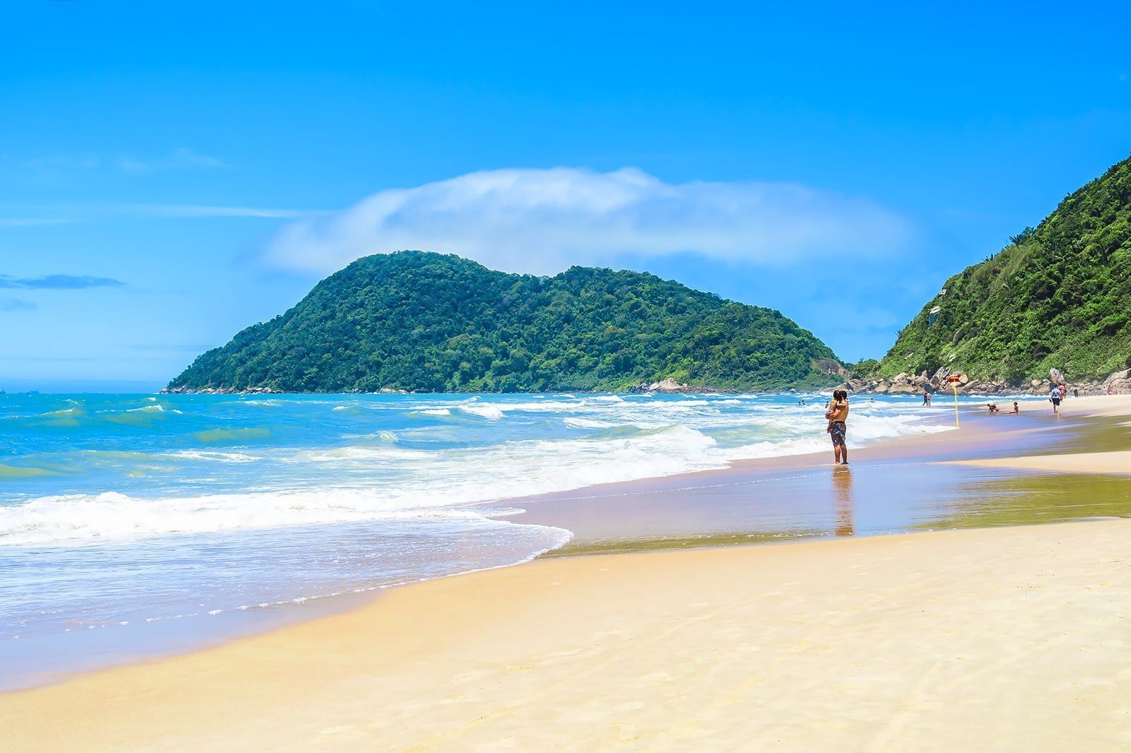 Sunny tropical beach with turquoise waves, green islands, and a person standing by the shoreline