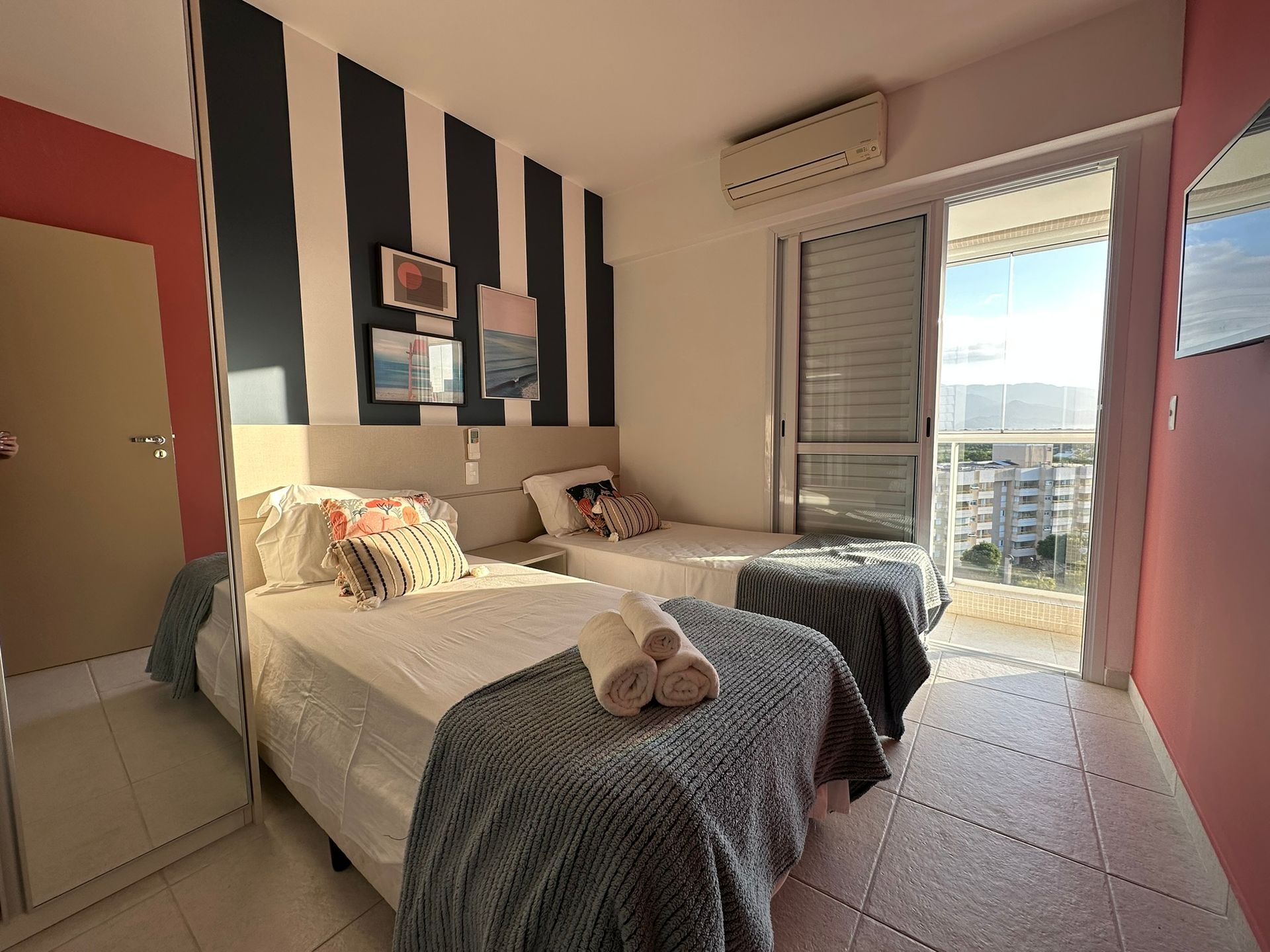 Quarto com duas camas de solteiro, parede com detalhes listrados, duas camas e acesso à varanda. Inclui roupa de cama em tons neutros e almofadas decorativas.