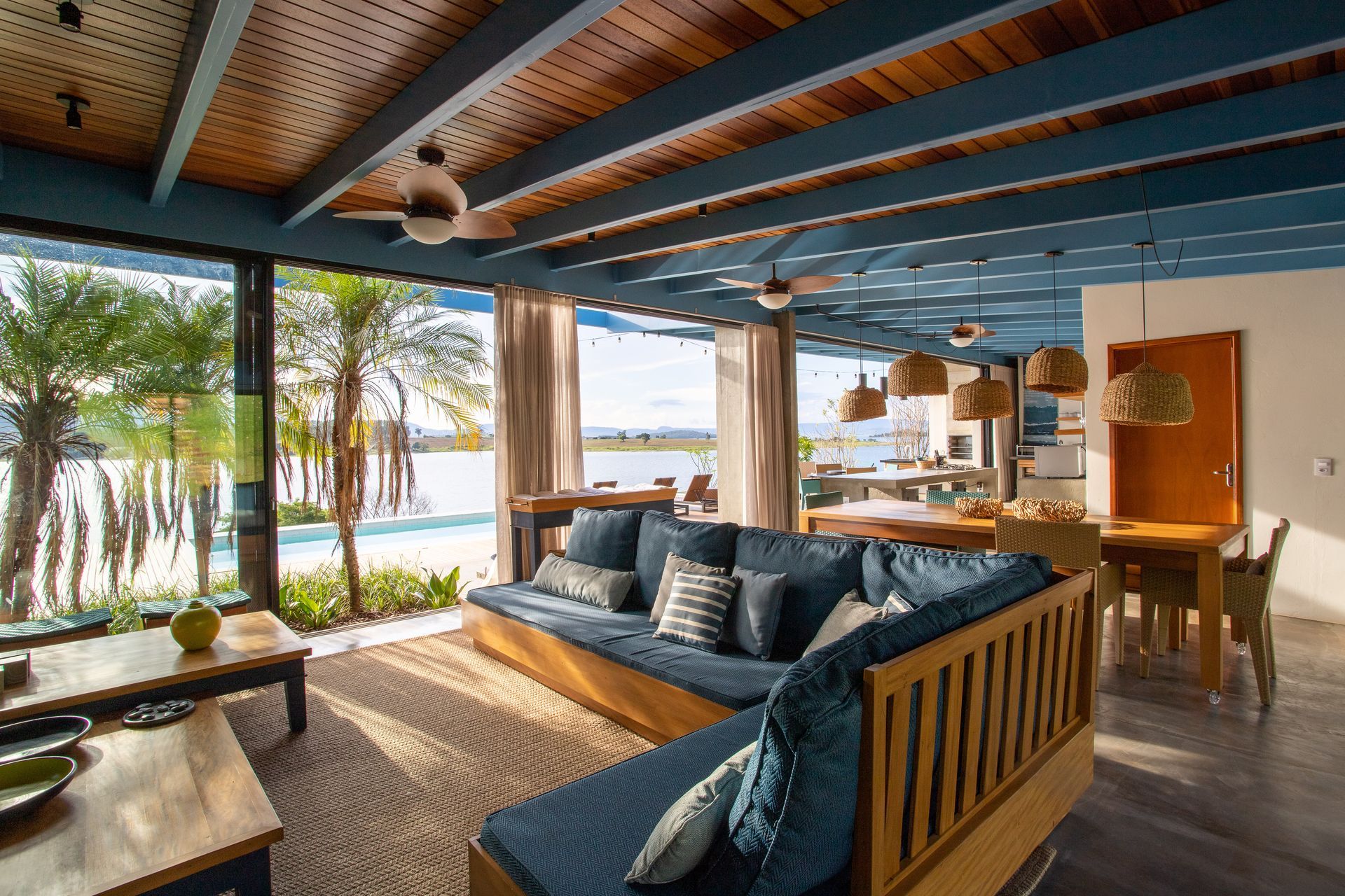 Uma sala de estar iluminada e aberta com vista para a praia. Sofás azuis, teto de madeira e janelas amplas preenchem o espaço.