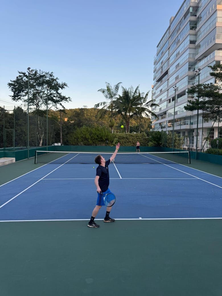 Uma pessoa saca uma bola de tênis em uma quadra azul, perto de um prédio alto, sob um céu limpo.