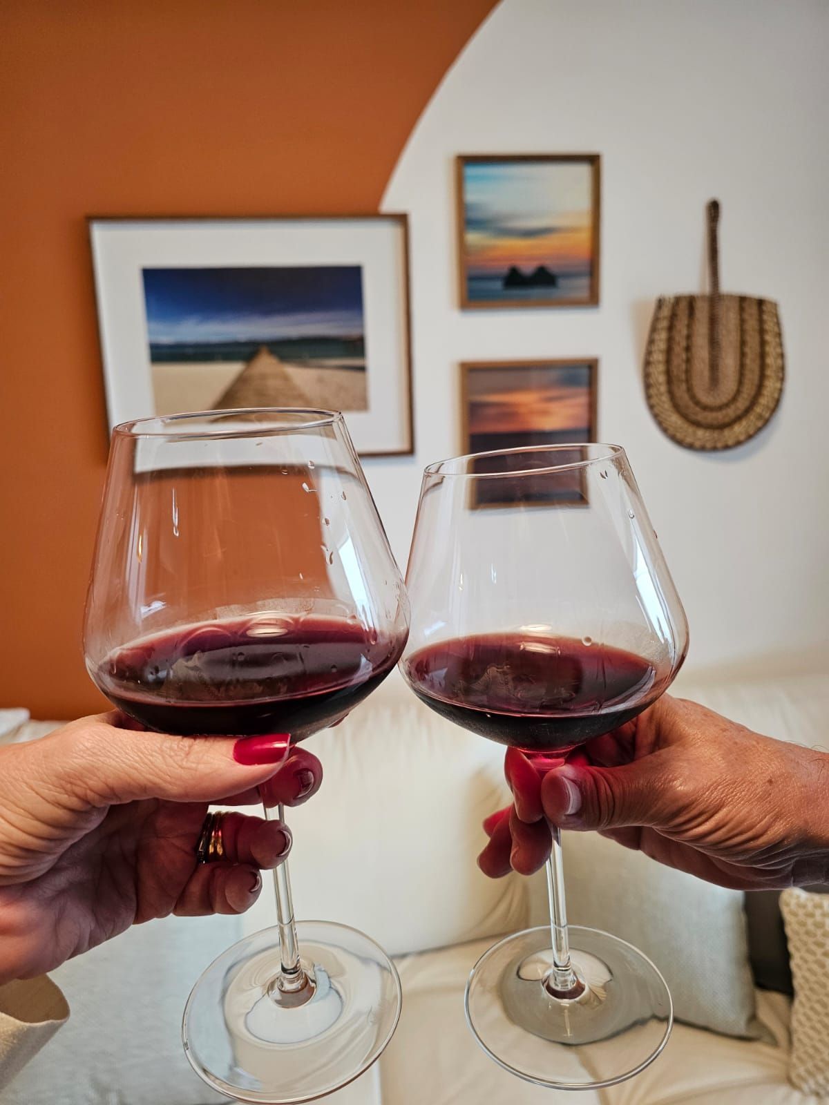 Duas mãos tilintando taças de vinho tinto em frente a uma parede com arte.