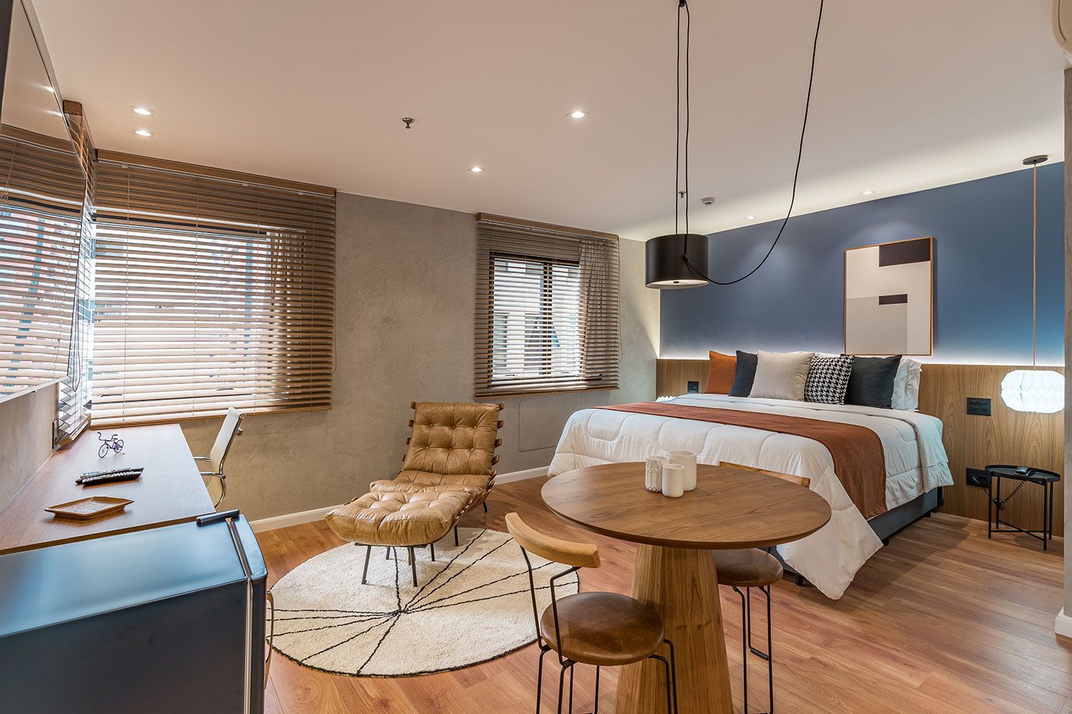 Quarto de hotel moderno com cama, mesa redonda com bancos, poltrona e janelas com persianas de madeira.