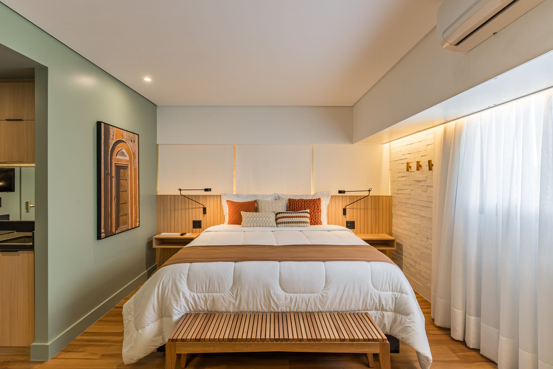Quarto com cama, banco, criados-mudos e obras de arte. As paredes são pintadas de verde e branco, com piso de madeira.