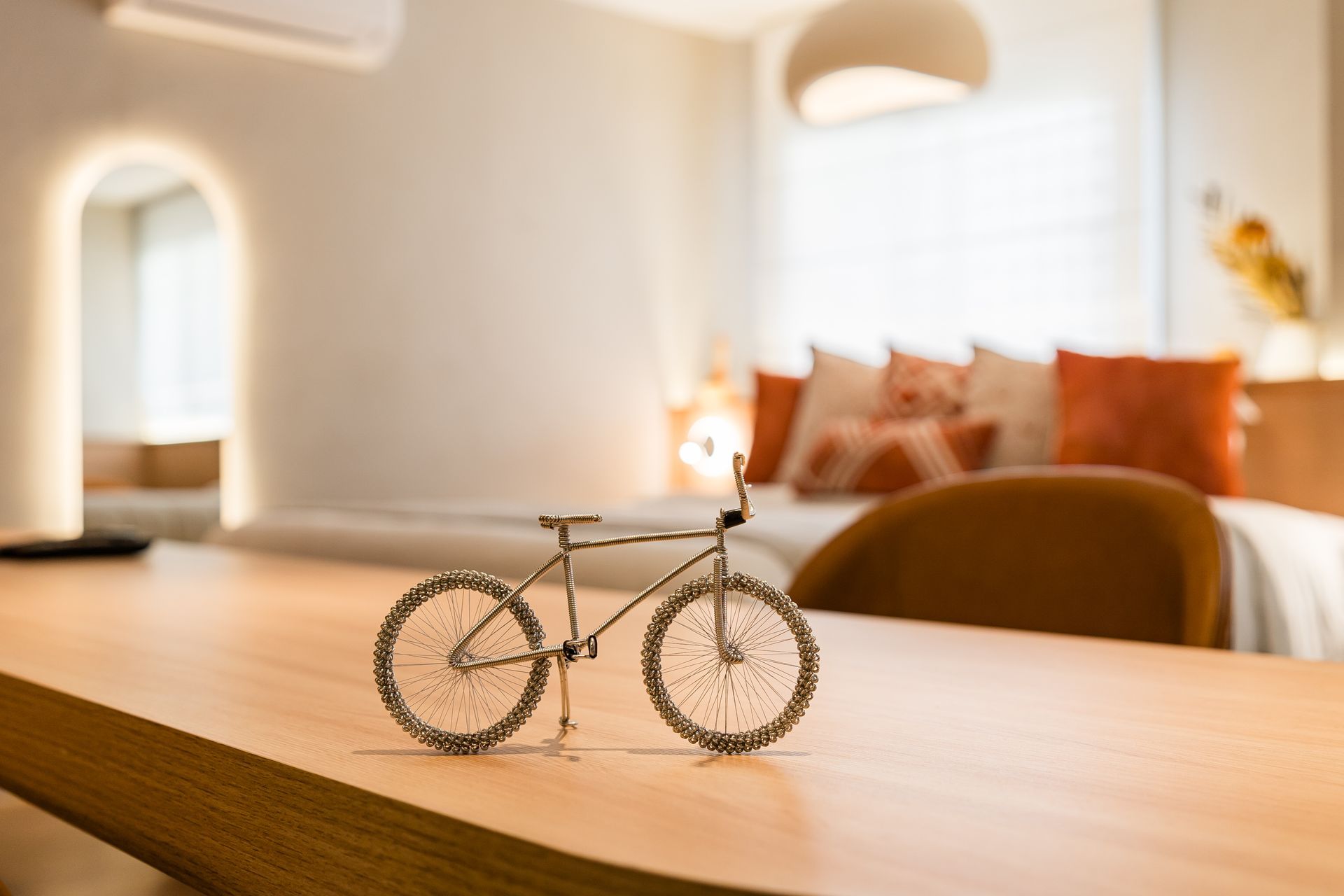 Uma pequena bicicleta de arame repousa sobre uma mesa de madeira clara em um quarto moderno e iluminado. Almofadas em tons de laranja adornam uma cama ao fundo.