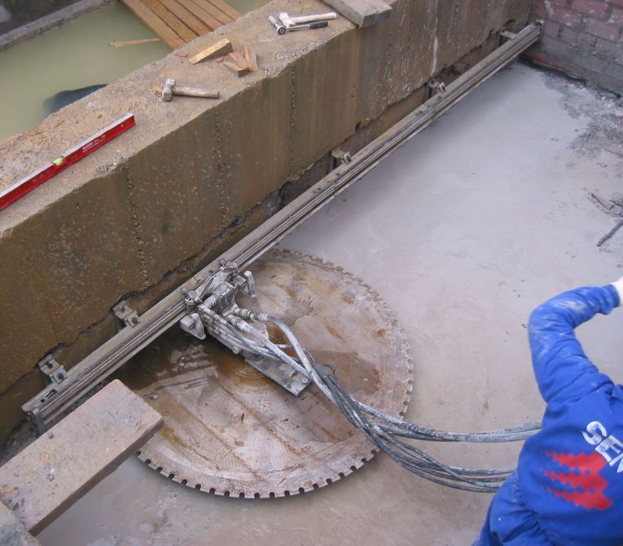 Una sierra para hormigón corta una losa junto a una pared, con un trabajador con equipo de protección azul