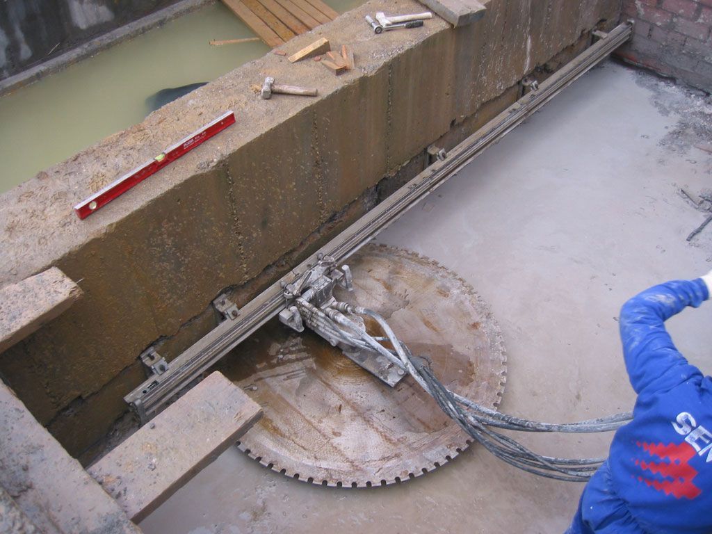 Una sierra circular cortando hormigón a lo largo de una pared, con chorro de agua y cables en una obra en construcción.