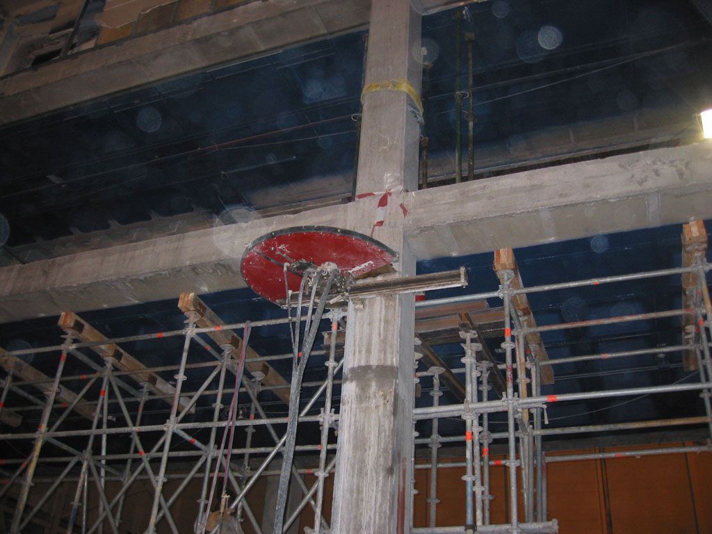 Andamio de construcción bajo un techo de hormigón, con un cubo o tapa roja colgando cerca de un pilar.