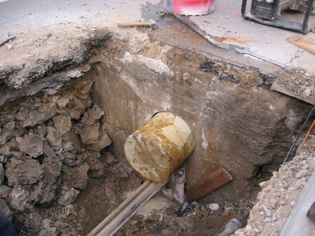 Zanja de construcción con tubería expuesta y cubo de hormigón en una excavación de tierra.