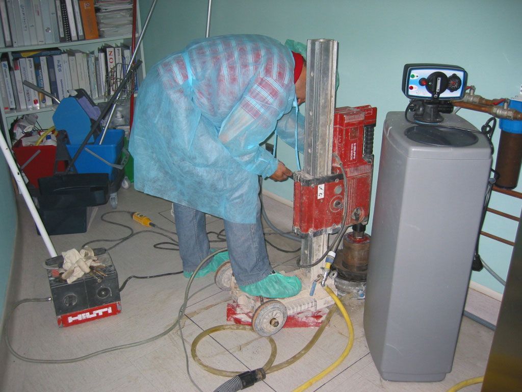 Una persona taladra el suelo con una herramienta eléctrica en una habitación desordenada, con cables y equipos cerca.