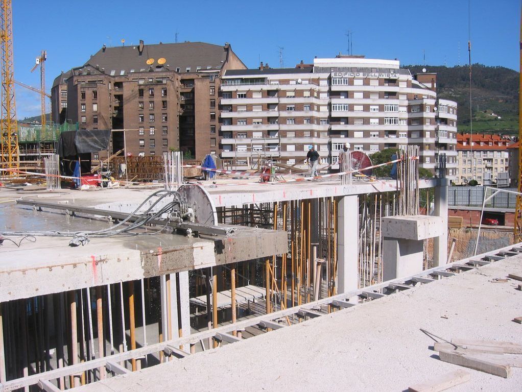 Obra en construcción con edificios de hormigón sin terminar y bloques de apartamentos bajo un cielo azul despejado.