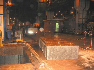 Obra en construcción nocturna y húmeda con trabajadores, barreras y bloques de hormigón bajo luces naranjas.