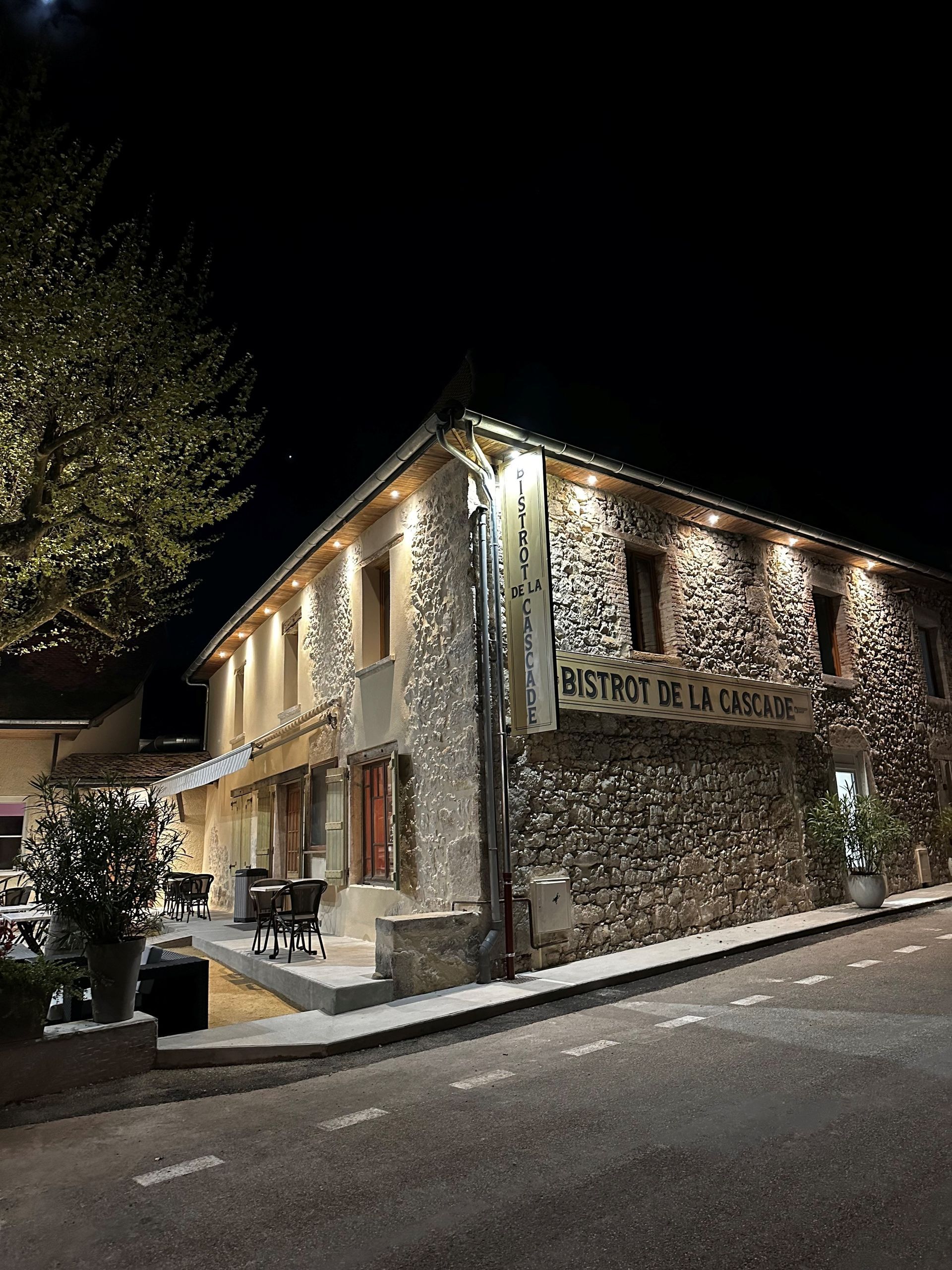 Bâtiment en pierre la nuit, éclairé par le bas. Enseigne du restaurant et tables en terrasse. Rue sombre.