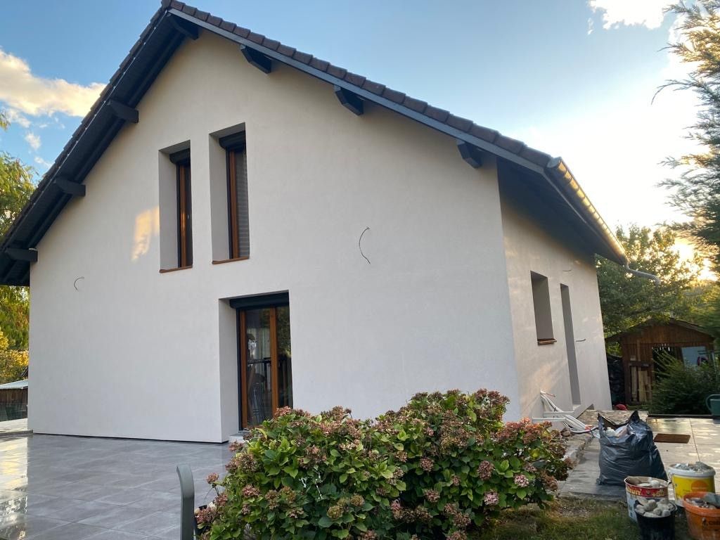 Maison beige avec un toit foncé, des fenêtres marron et une terrasse en béton.